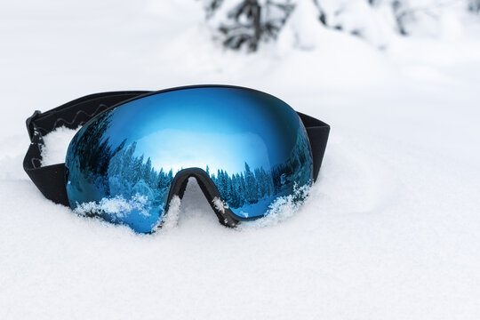 Ski goggles with blue mirrored lens lying on fresh snow, reflecting winter forest and mountains, symbolizing winter sports and adventure, Christmas holidays and ski vacation concept