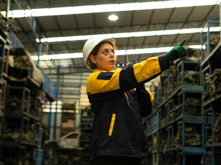 Hispanic female foreman worker holding walkie talkie, checking inventory of auto parts and engine maintenance duties in factory.