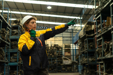 Hispanic female foreman worker holding walkie talkie, checking inventory of auto parts and engine maintenance duties in factory.