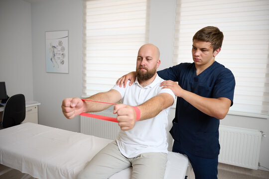 Physiotherapist guiding male patient with resistance band exercise for shoulder rehabilitation in clinic. Physical therapy session focused on injury recovery, strengthening, mobility improvement
