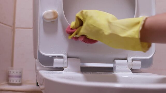 Hand in yellow gloves scrubs and clean the toilet hinge with a sponge, removing stains and residue