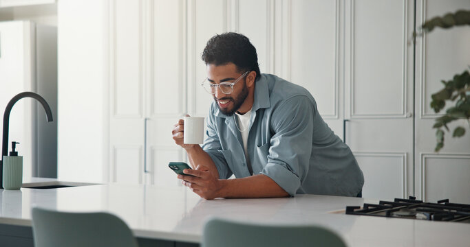 Phone, coffee and man in kitchen in home with networking, communication or social media on mobile app. Tea, technology and male person with cellphone for contact, texting or chatting in apartment.