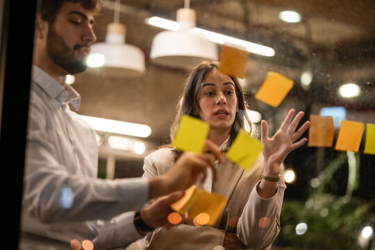 Colleagues brainstorming ideas on glass wall with sticky notes