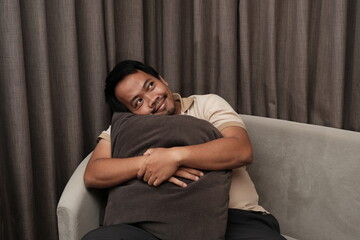 Southeast Asian man in a beige shirt sitting on a sofa, hugging a dark pillow while smiling and gazing upward with a dreamy expression.