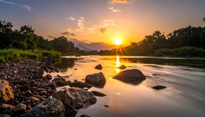 A serene river flows towards a brilliant sunset, framed by lush trees and mountainous terrain. The water mirrors the vibrant orange and golden hues