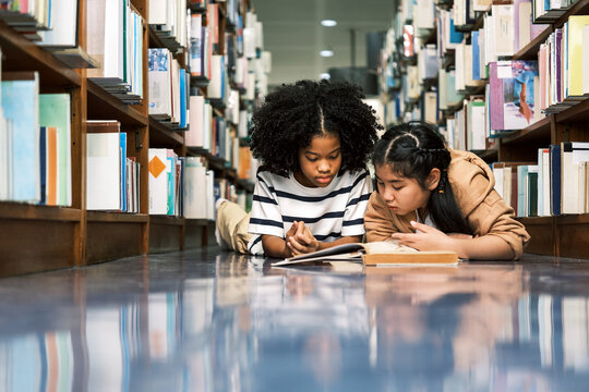 Two diverse children sit on the floor between bookshelves in a library, reading and sharing a book. A joyful academic moment filled with kids literature, knowledge, and classroom curiosity.