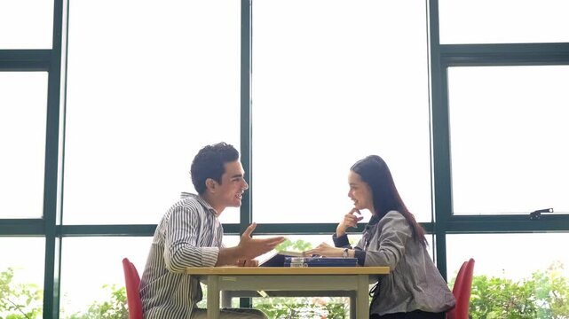 Two people sit in the library across a table by large windows, engaged in a friendly discussion with books on the desk, suggesting study or a meeting in a bright learning space.