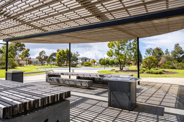 A contemporary open-air park shelter with built-in barbecue facilities, picnic tables, and a slatted louvered roof for shade in an Australian suburban park. Recreational amenities in outdoor space.