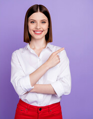 A happy young woman with short brown hair, wearing a white shirt and red pants, smiling and pointing her index finger to the side against a purple background.