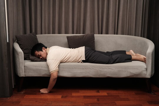 Southeast Asian man in casual clothes lying face down on a grey sofa, falling asleep, appearing exhausted or overwhelmed.