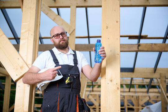 Construction worker taking a break and drinking water at wooden house frame construction site. Builder resting with hard hat and water bottle. - Powered by Adobe