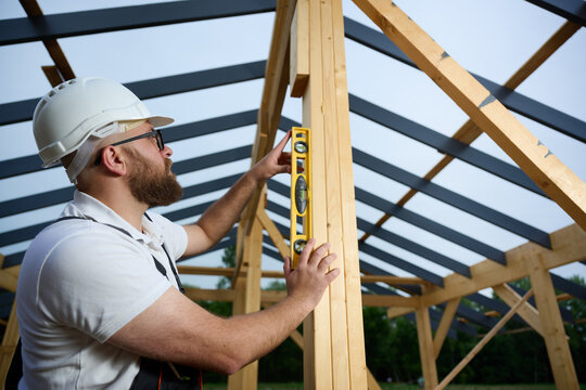 Construction worker using spirit level to check vertical alignment of wooden beam at frame house site. Builder wearing hard hat and safety glasses working on wooden structure outdoors.