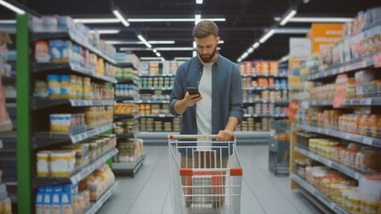Man using smartphone while pushing shopping cart down grocery store aisle - Powered by Adobe