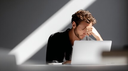 Stressed young man holds his head working on a laptop at a desk with strong sunlight and shadows