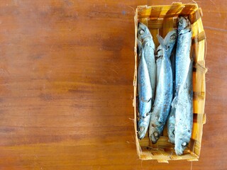 A woven basket holds several small, cooked or dried fish with silver and blue hues, resting on a warm wooden surface. Ikan pindang from a local market in Malang, Indonesia.