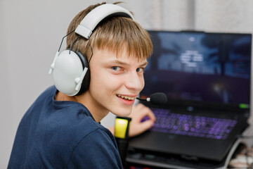 Happy smiling teenage boy wearing a headset with a microphone, turning to look at the camera while enthusiastically playing a video game on a laptop