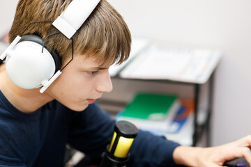 Focused young male gamer or streamer wearing large white headphones, working on a laptop with a professional condenser microphone nearby