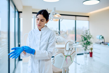 Fototapeta premium Female dentist or assistant preparing for work, putting on blue protective gloves in a modern dental clinic or office