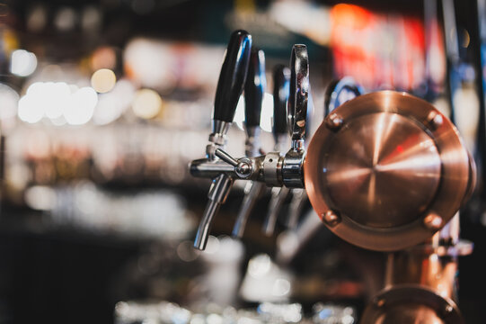 Close-up, selective focus shot of several modern, shiny beer taps with black handles on a bar counter in a dimly lit pub or restaurant setting