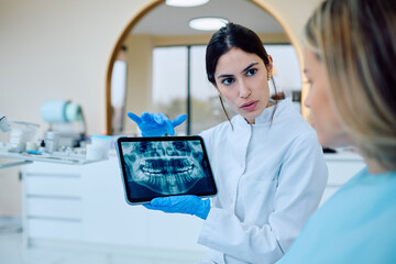 Dentist in white coat and blue gloves showing a panoramic dental x-ray on a tablet to a female patient in a dental clinic