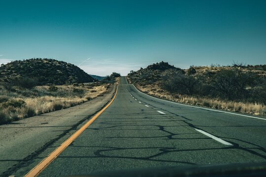 Empty cracked asphalt road stretching through a dry desert landscape under a clear blue sky with distant hills