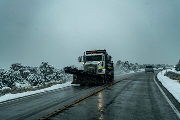 Snow plow truck clearing a wet road during a snowy winter day on a rural highway surrounded by snow-covered vegetation