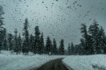 Blurred snowy road view through a rain-covered window with evergreen trees lining the roadside under cloudy sky