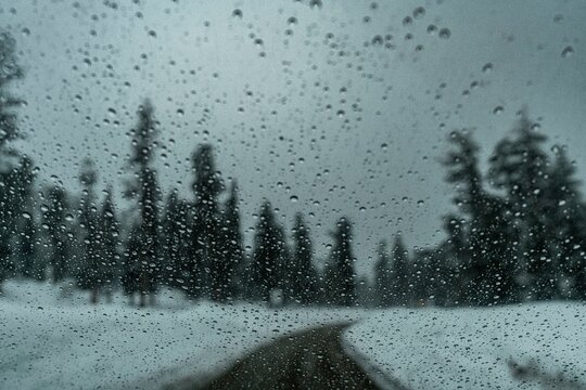 Blurred snowy road view through a rain-covered window with evergreen trees lining the roadside under cloudy sky - Powered by Adobe