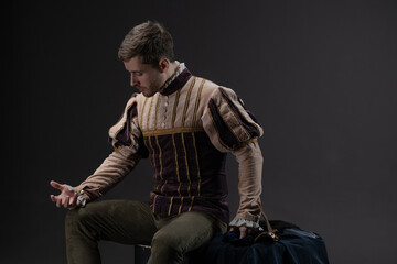 Aristocratic character from early Renaissance period dressed in elaborate brocade fabric with cape accessory and beret, captured in dramatic stance on gray backdrop