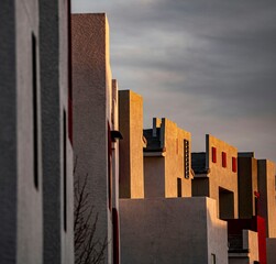 Modern adobe-style buildings with warm sunlight casting shadows on textured walls under a dramatic cloudy sky