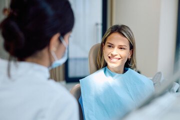 Young woman sitting in a dental chair smiling at a dentist, discussing her oral hygiene and receiving a checkup