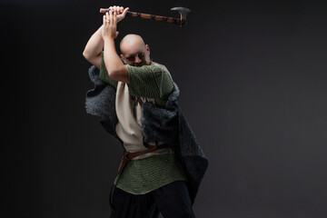 Medieval Viking warrior portrayal: bald man with weapons and fur on neutral gray, He swung his axe to strike