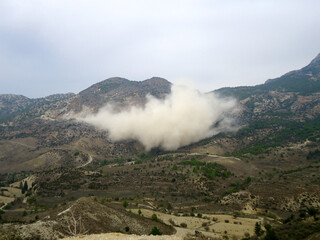 A massive cloud of white dust and fumes hanging low over a shrub-covered hillside following an explosion or blasting at a quarry, illustrating environmental disturbance and mining effects.