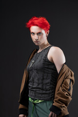 A bold young woman with vibrant red hair wears oversized acid green trousers and an aviator jacket with modern glasses, posing against a plain gray studio backdrop