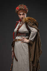A young Viking woman, a combat heroine in a fantasy Nordic costume with a Scandinavian battle axe in her hands, a natural-colored dress and a cape with fur, photo on gray