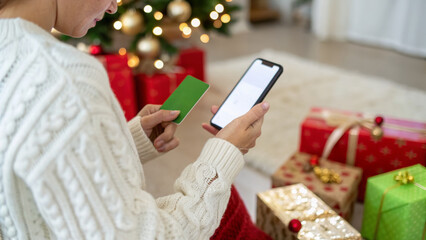 Young person in cozy sweater using smartphone and credit card for holiday smart spending during Christmas and new year gift shopping with wrapped presents and tree lights