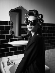 Stylish woman in hair rollers and oversized sunglasses poses in a retro black & white bathroom, capturing a chic, vintage beauty routine moment. Perfect for fashion, beauty, or lifestyle themes.