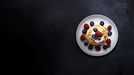 A top-down view of golden pancakes topped with fresh berries and mint on a light plate against black