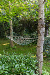 Green netted hammock suspended between two trees in lush garden, surrounded by greenery and dappled sunlight.