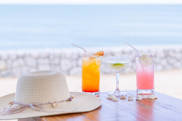 Summer beach scene with three colorful cocktails on table, straw hat, seashells. Relaxing ocean view, concept of leisure, tropical vacation, refreshing drinks by sea.