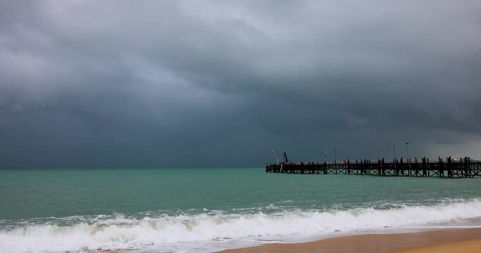 Tropical beach with turquoise water and a wooden pier under dramatic storm clouds. The approaching rain and dark sky contrast with the bright sea, creating a cinematic and moody coastal atmosphere.
