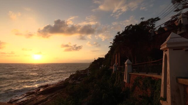 Tropical seaside timelapse showing a golden sunset over the ocean with waves crashing against the rocky shore and warm light illuminating the coastal cliffs and architecture. A serene, cinematic scene