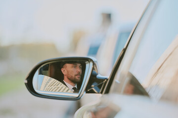 Caucasian young adult man wearing suit and tie reflected in car side mirror driving vehicle outdoors, facial expression serious and focused, short beard visible