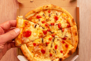 Hand holding slice of fresh pizza above open delivery box, showing melted cheese and tomato sauce, concept of fast food and casual dining