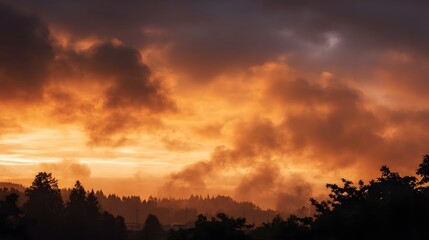 Fototapeta premium Dramatic sky at dawn or dusk with vibrant orange and red clouds illuminating a silhouetted forest and hills