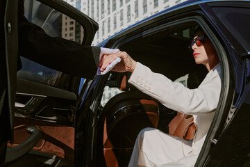 Caucasian middle aged woman exiting car while holding hand of man in suit, wearing sunglasses, business district buildings visible in background, formal interaction occurring
