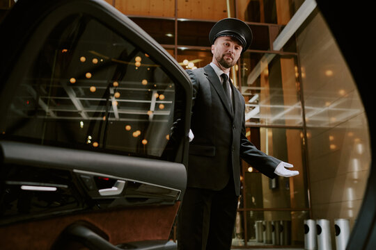 Caucasian middle aged man wearing uniform and cap opening car door with gloved hand, standing in modern building entrance, looking at camera, providing professional chauffeur service