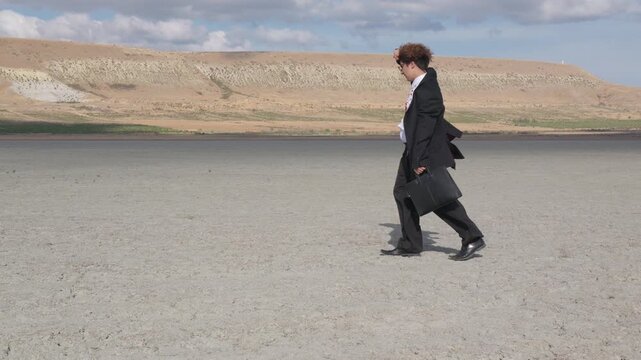 A curly-haired teenager with glasses and a mature business suit stubbornly walks forward, shielding himself from the wind with his hand, against the backdrop of a hill and blue sky