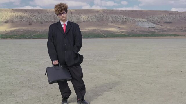 A sad teenager in a mature business suit with a briefcase stands on dry ground against the backdrop of a hill with a vineyard. The camera zooms out