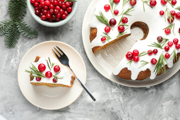 Delicious cut Christmas cake with icing, cranberries, rosemary and fir tree branches on grey textured table, flat lay